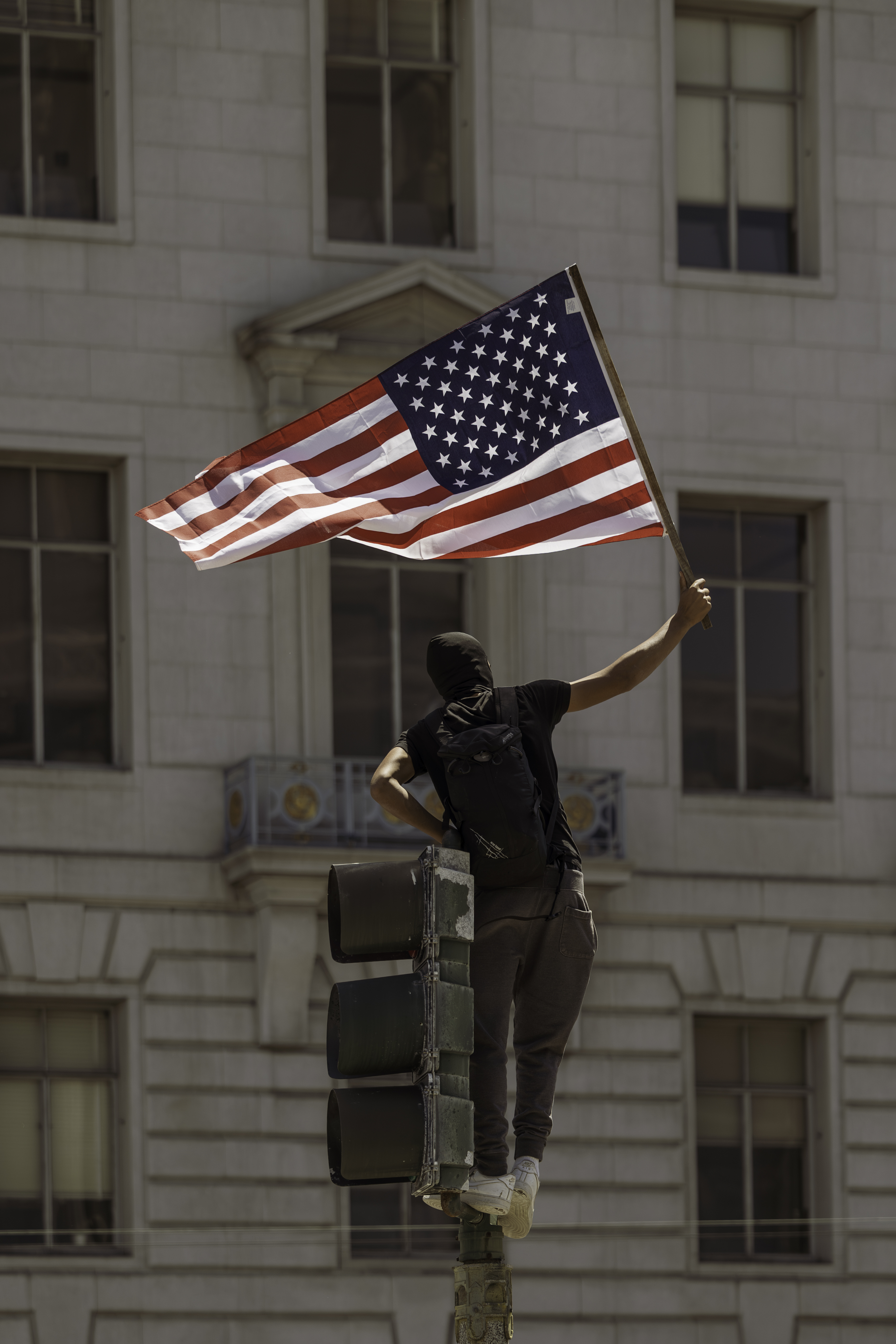 Masked figure waving American flag atop a traffic light, San Francisco protest