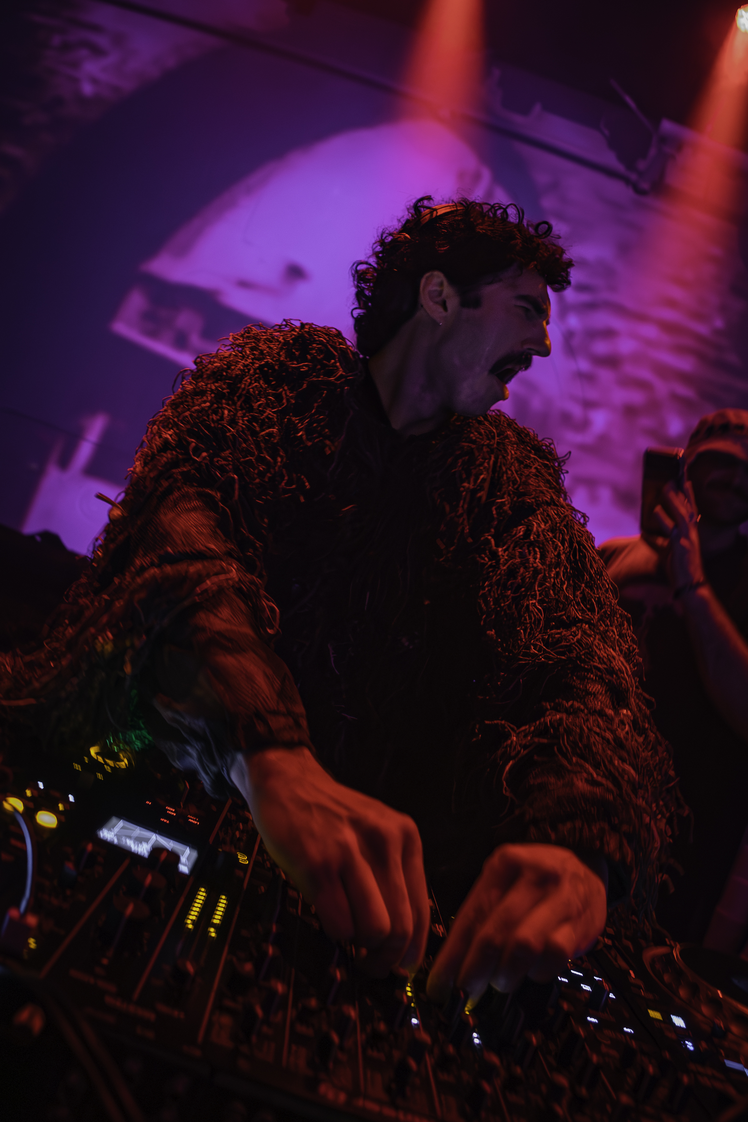 DJ in elaborate fringe costume leaning over decks under purple and red club lights