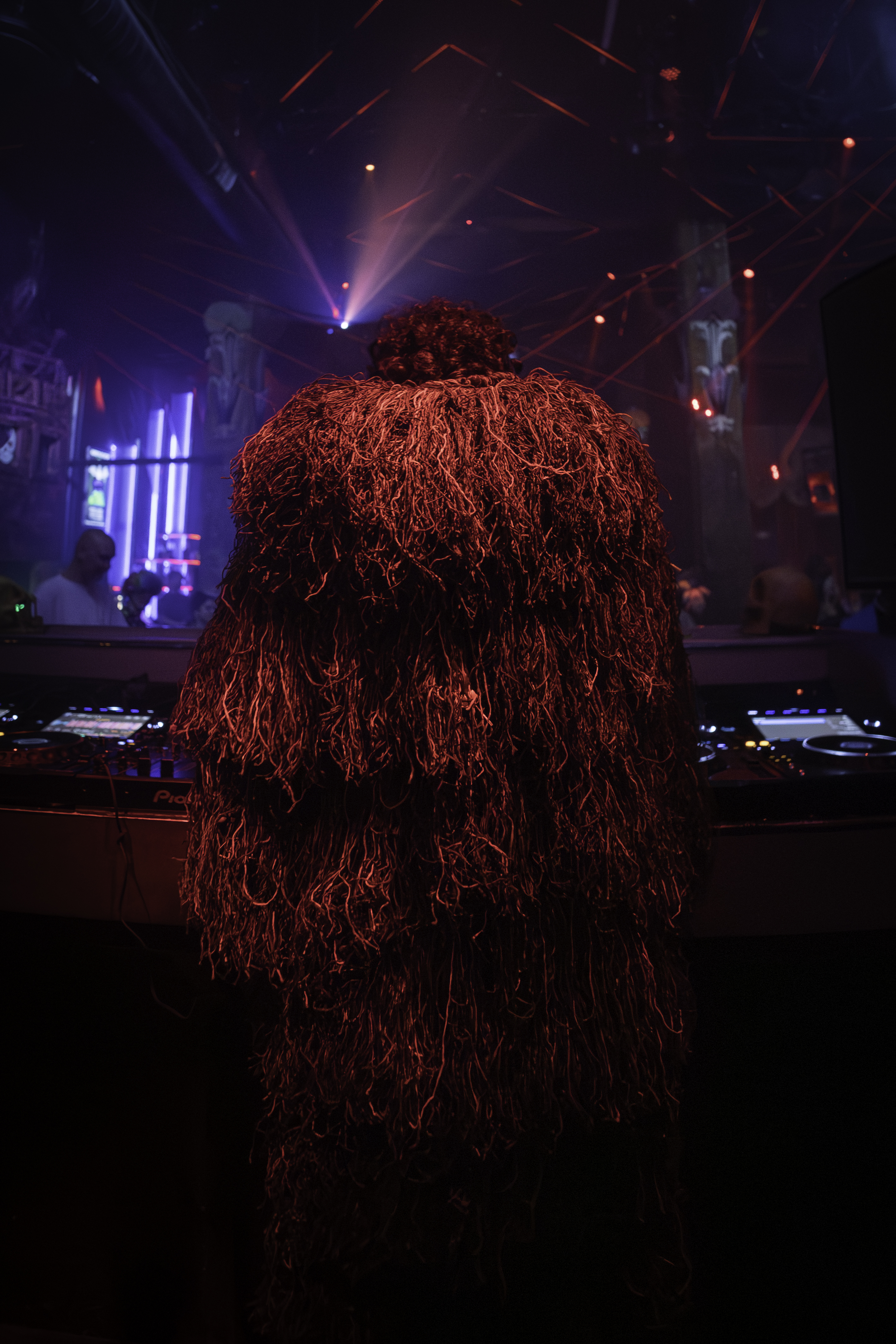 DJ in shaggy textured costume viewed from behind, laser lights cutting through venue